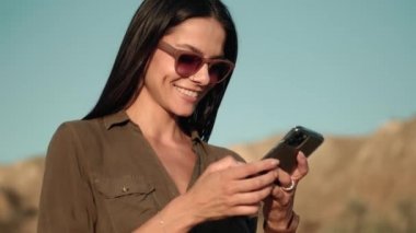 Güneş gözlüğü takan gülümseyen bir kadın yazın dışarıda yürürken cep telefonunu kullanıyor.