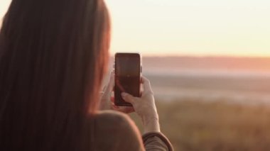 Yazın gün batımında akıllı telefonuyla manzara fotoğrafı çeken bir kadın görülüyor.
