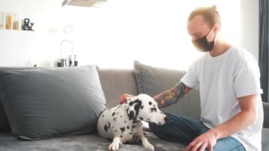 A man wearing protective medical mask while sitting and petting his dalmatian dog at home