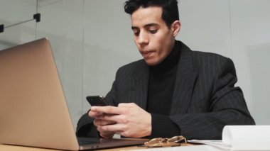 Yakışıklı bir Arap adam ofiste otururken akıllı telefonunu kullanıyor.