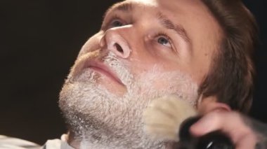 Yakından bakınca, berber dükkanında saç fırçasıyla yakışıklı, yetenekli bir berber müşterinin yüzünü, saç kesiminden sonra yüzünü temizliyor.
