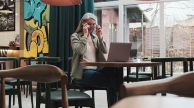 Memnun bir kadın içerideki kafede otururken bilgisayarını kullanarak gözlük takıyor.