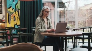 Gözlüklü ciddi bir kadın içerideki kafede otururken bilgisayarını kullanıyor.