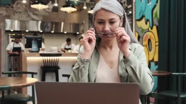 Gülümseyen bir kadın içerideki kafede otururken bilgisayarına bakarken gözlük takıyor.