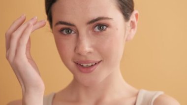 Mutlu güzel bir kadının yakın çekim görüntüsü stüdyodaki bej duvarın üzerinde izole bir şekilde kameraya poz veriyor ve göz kırpıyor.