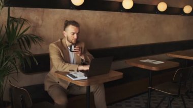 Ceket giyen yakışıklı beyaz bir işadamı kafede kahve içerken bilgisayarını kullanarak çalışıyor.