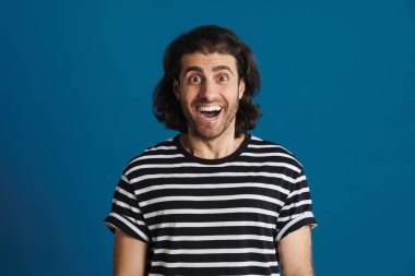 Çizgili tişörtlü heyecanlı esmer adam mavi arka planda izole edilmiş kameraya bakıyor.