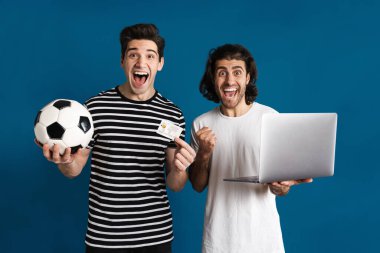 Futbol ve kredi kartını mavi arka planda tutarken dizüstü bilgisayar kullanan iki adam heyecanlandı