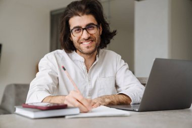 Gülümseyen orta yaşlı esmer Türk adam evde dizüstü bilgisayarla oturmuş notlar yazıyor.