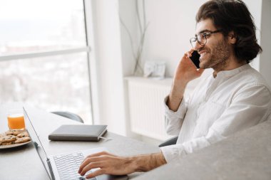 Gülümseyen orta yaşlı esmer Türk adam evde dizüstü bilgisayarla oturmuş cep telefonuyla konuşuyor.