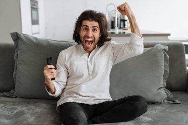 Evdeki kanepede oturup kutlama yaparken elinde uzaktan kumandayla mutlu bir genç adam.