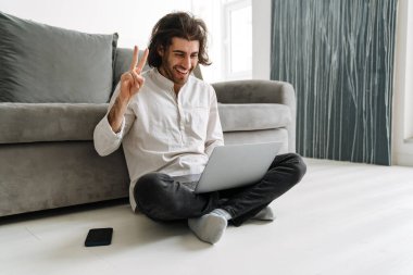 Orta yaşlı, esmer, Türk bir adam evde dizüstü bilgisayarla oturmuş el sallıyor.
