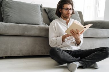 Orta yaşlı, esmer, Türk bir adam oturma odasında oturmuş kitap okuyor.