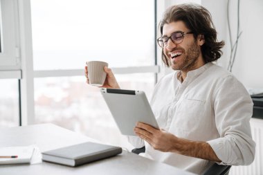 Beyaz gömlekli, orta yaşlı, esmer, Türk bir adam masada oturmuş elinde kupayla tablet bilgisayarına bakıyor.