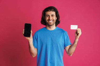 Tıraşsız, tişörtlü, kredi kartı ve akıllı telefonu pembe arka planda gösteren bir adam.