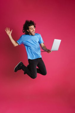 Heyecanlı esmer adam pembe arka planda izole bir şekilde sıçrarken diz üstü bilgisayar kullanıyor.