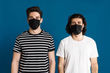 Beyaz esmer, maskeli iki adam mavi arka planda izole edilmiş kameraya bakıyor.