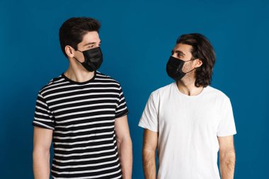 Beyaz esmer, maske takmış iki adam mavi arka planda izole bir şekilde birbirlerine bakıyorlar.