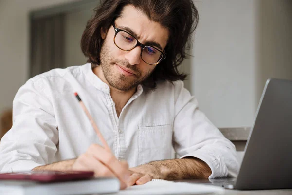 闷闷不乐的中年黑发男人坐在桌旁 家里带着笔记本电脑在写便条 — 图库照片
