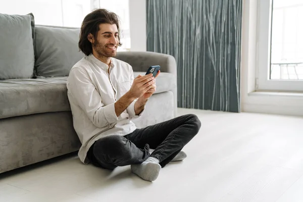 Smiling mid aged turkish man holding mobile phone sitting on a floor in the living room on a video call