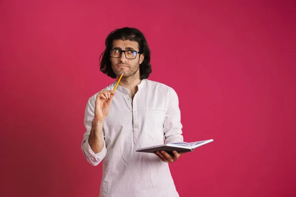 White puzzled man in eyeglasses frowning and writing down notes isolated over pink background