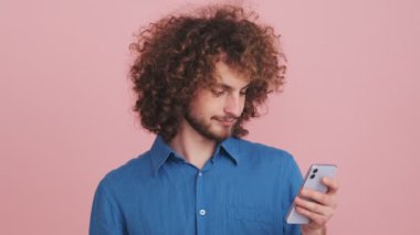 Yakışıklı bir kıvırcık adam telefonuyla stüdyoda pembe arka planda duruyor.