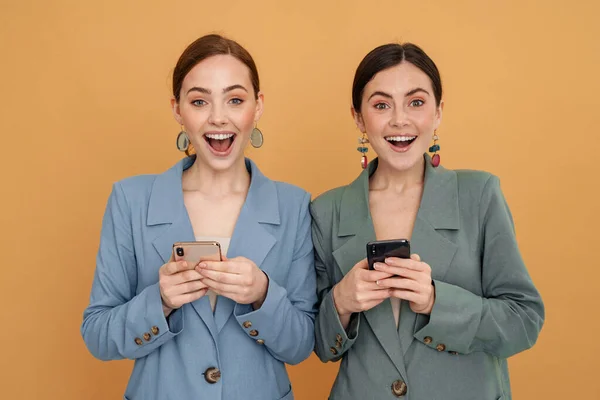 黄色の背景に隔離された携帯電話を使用している間に笑みを浮かべて若い2人の女性 — ストック写真