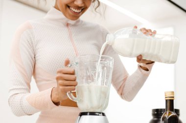 Siyahi genç kadın gülümsüyor ve evde sütle smoothie yapıyor.
