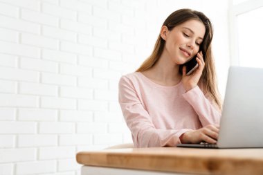 Lounge giysileri giyen mutlu beyaz bir kadın masada dizüstü bilgisayarla oturuyor, çalışıyor, cep telefonuyla arıyor.