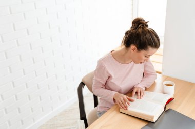 Salondaki genç beyaz bir kadının en iyi görüntüsü evdeki masada oturmuş kitap okuyor.