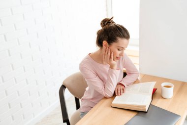 Salondaki gülümseyen beyaz bir kadının en üst görüntüsü evdeki masada oturmuş kitap okuyor.