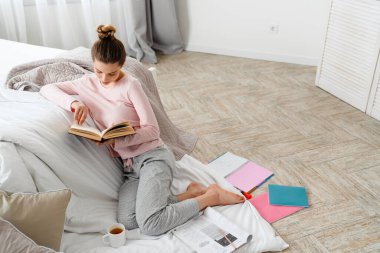 Genç beyaz kadın kitap okuyor. Yatak odasının zemininde, battaniyenin üzerinde oturuyor.