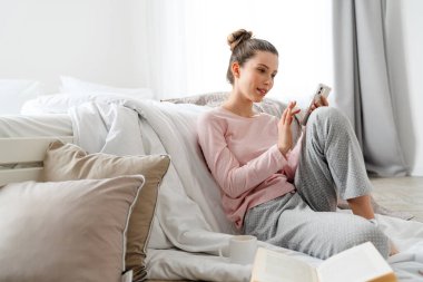 Salondaki gülümseyen beyaz kadın, elinde cep telefonuyla yatak odasında yerde oturuyor.