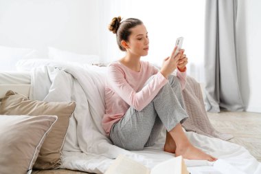 Salondaki gülümseyen beyaz kadın, elinde cep telefonuyla yatak odasında yerde oturuyor.