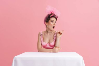 Pembe arka planda soyutlanmış bir masada otururken parmağını kenara çeken şaşkın kadın.