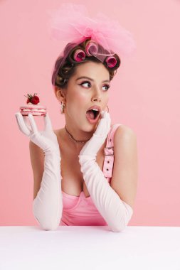 Şok olmuş bir kadın, pembe arka planda tek başına oturmuş pasta yerken eldiven takıyor.