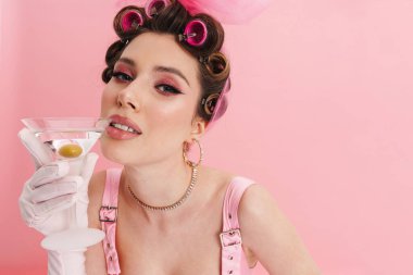 Pembe elbiseli genç beyaz kadın barbi pembe duvarın üzerinde duruyor, elinde kokteyl var.
