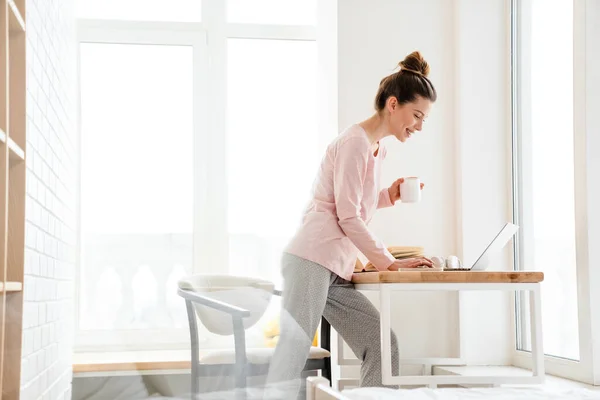 ラウンジの服を着た幸せな若い白人女性ラップトップコンピュータでテーブルに立って ビデオ通話 マグカップを保持 — ストック写真