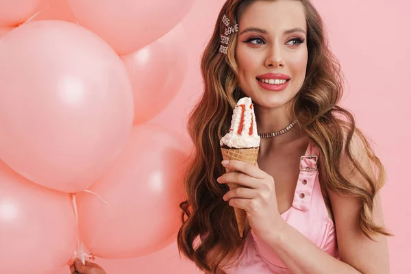 年轻的微笑的女人一边吃着冰激凌 一边拿着粉色背景的气球摆姿势 — 图库照片