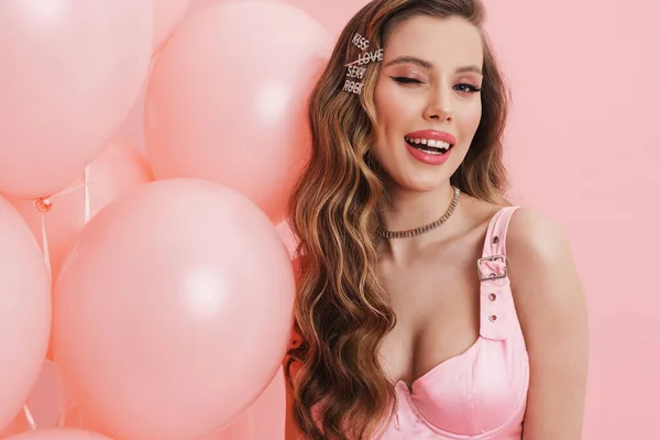 年轻的微笑的女人 穿着连衣裙 与粉色背景的气球一起摆姿势时眨眼 — 图库照片