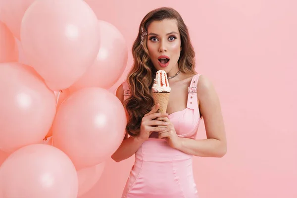 年轻而震惊的女人一边吃着冰激凌 一边拿着粉色背景的气球摆姿势 — 图库照片