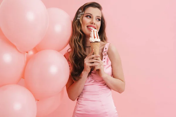 年轻的微笑的女人一边吃着冰激凌 一边拿着粉色背景的气球摆姿势 — 图库照片