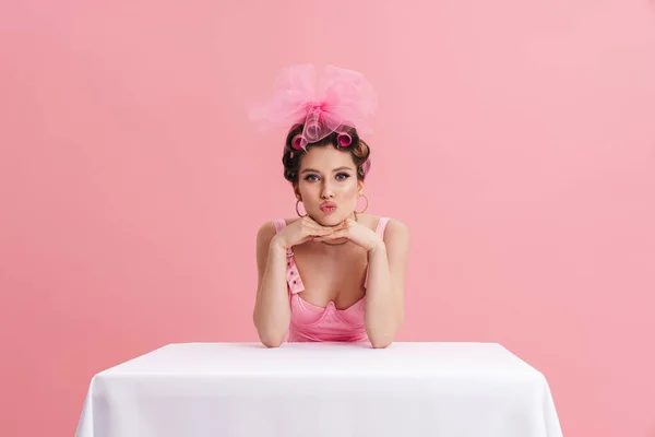 Молодая Женщина Бигудях Целует Губы Сидя Столом Изолированы Розовом Фоне — стоковое фото