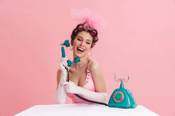 年轻女子戴着手套 摆出复古式电话的姿势 坐在被粉色背景隔开的桌子旁 — 图库照片