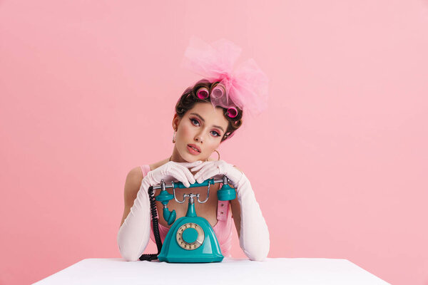 Young woman wearing gloves posing with retro phone while sitting at table isolated over pink background