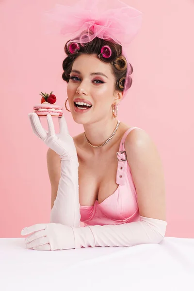 微笑的女人戴着手套吃蛋糕 坐在餐桌旁 与粉红的背景隔离 — 图库照片