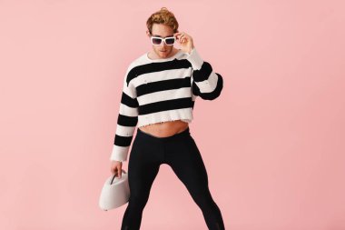 Kızıl saçlı, güneş gözlüklü genç adam pembe duvarda izole edilmiş halterli poz veriyor.