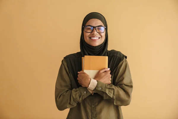 Black muslim woman in eyeglasses smiling while posing with book isolated over beige background