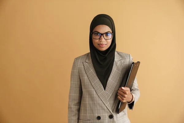 戴着眼镜的黑人穆斯林妇女摆出与米色背景隔离的剪贴板的姿势 — 图库照片