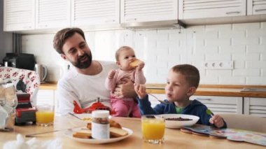 Mutlu bir baba genç kızını kucağında tutarken oğlu mutfakta müsli yiyor.